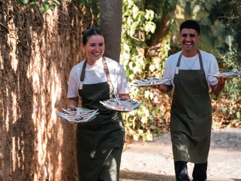 Dos camareros de Grupo Picolín sonriendo mientras llevan bandejas de aperitivos en una finca
